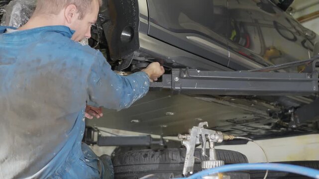 Man sitting beneath car on lift removes cap, inserts spray gun tube into sill cavity, applying lanolin rustproofing