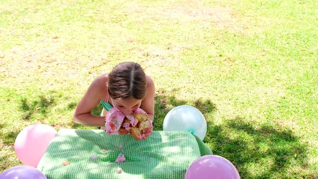 Loony birthday girl eating cake with bare hands on grass outdoors, slomo