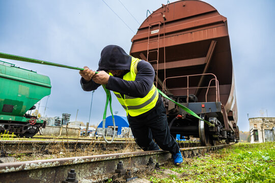 A hooded man in a high-visibility vest strains to pull a heavy freight wagon using a green rope along railway tracks in an industrial yard
