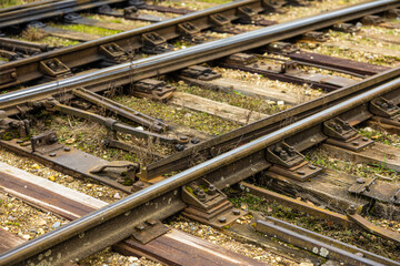 Fototapeta premium Detailed view of a railway track switch with metal components, bolts, and overgrown vegetation between the tracks. Weathered wood and gravel visible throughout