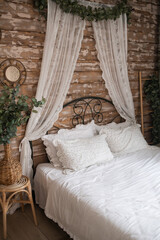 Details of bright elegant interior in cozy bedroom. Wicker side table with vase and flowers next to comfortable bed with white linen bedding. 