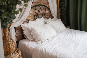 Details of bright elegant interior in cozy bedroom. Wicker side table with vase and flowers next to comfortable bed with white linen bedding. 