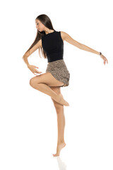Obraz premium Studio shot of tall young ballerina with long brown hair in black top and leopard mini skirt posing gracefully on tiptoes full body on white background.