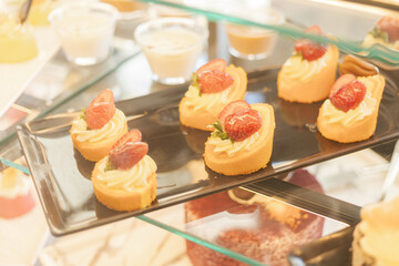 A selection of fresh, cream-filled strawberry desserts is beautifully arranged on a black tray in a bakery. The afternoon light accentuates the vibrant colors of the treats