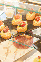 Assorted pastries topped with fresh strawberries are beautifully arranged on a glass display stand in a pastry shop during daylight. The inviting colors and textures highlight the desserts