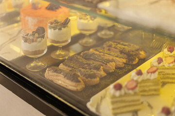 A variety of colorful pastries and desserts are beautifully arranged in a bakery display case. The scene captures treats like eclairs, layered cakes, and parfaits, inviting customers