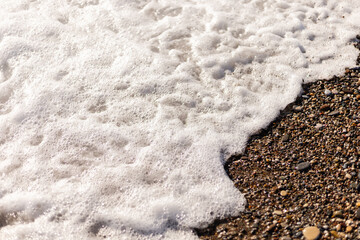 Gentle waves roll onto a sandy beach, creating a frothy foam along the shore. Small pebbles and grains of sand are visible as water recedes under the bright sun