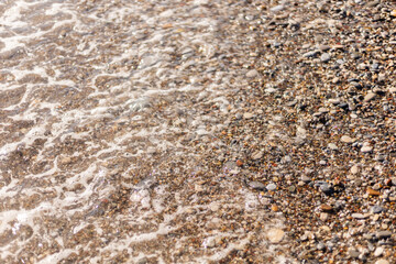 The shoreline features smooth pebbles and sand as gentle waves wash ashore. Sunlight glimmers on the water, highlighting the colors of the stones