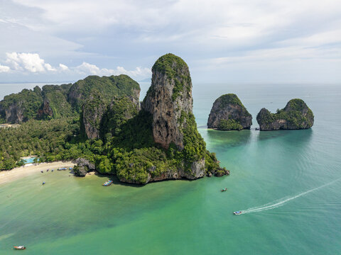 Aerial view of towering limestone cliffs draped in emerald foliage meet the turquoise waters, boats skimming the surface near the sandy shores, Ao Nang, Krabi, Thailand.