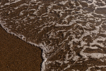 Soft waves roll in, creating foam on the shore as sunlight sparkles on the water. The sandy beach invites relaxation, with a peaceful atmosphere and warm weather