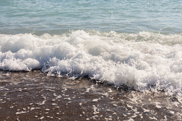 Gentle waves roll onto the shore, creating white foam as they reach the sandy beach. The sun reflects off the water, enhancing the serene summer atmosphere