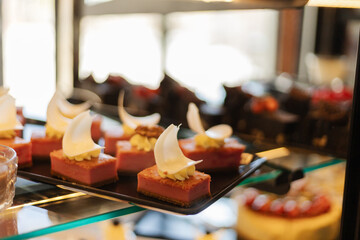 A selection of elegant mousse desserts sits on a glass display case, showcasing vibrant colors and creamy toppings. The cozy cafe ambiance adds warmth to the scene