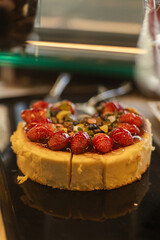 A beautifully crafted cheesecake sits on a black plate, adorned with vibrant red strawberries and colorful nuts on top. This dessert is showcased in a cafe environment, inviting patrons to indulge