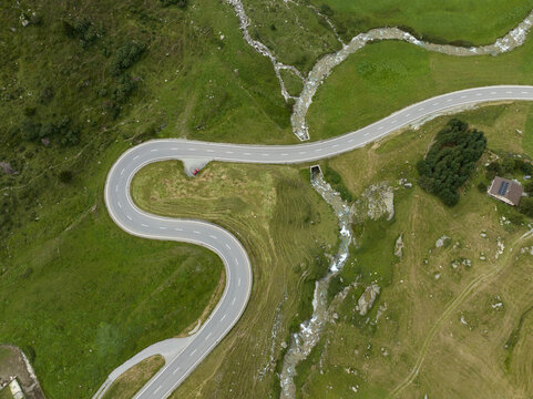 Aerial view of a winding road cutting through lush green meadows, alongside a stream, creating a striking contrast of natural and man-made landscapes, Surses, Grisons, Switzerland.