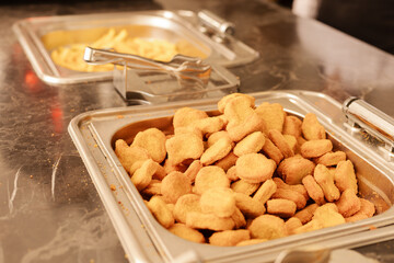 A large tray holds a mound of crispy chicken nuggets, freshly prepared, while another tray nearby features golden fries. This scene is set in a busy cafeteria during lunch