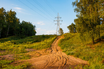 A winding dirt path ascends a grassy hill towards a towering electricity pylon under a bright summer sky. Lush greenery borders the rustic route