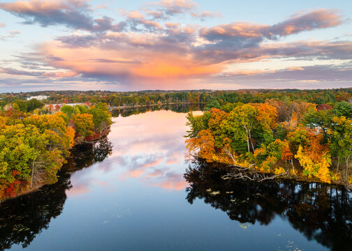 Aerial view of Fisk Pond reflecting the vibrant hues of autumn foliage under a dramatic sunset. Natick, Massachusetts, USA.