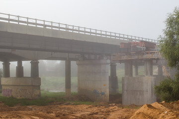 A sturdy concrete bridge under renovation emerges from a dense, hazy morning fog, its supports...