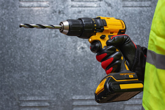 Close-up of a worker holding a yellow cordless drill with a drill bit, wearing protective gloves and safety vest. Concept of construction, power tools, and professional craftsmanship.