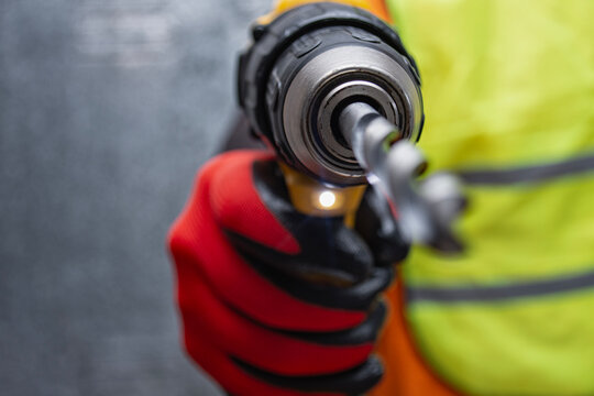 Close-up of a worker holding a yellow cordless drill with a drill bit, wearing protective gloves and safety vest. Concept of construction, power tools, and professional craftsmanship. - Powered by Adobe