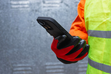 Construction worker wearing protective gloves and reflective safety vest while holding a smartphone. Concept of modern industrial communication, safety at work, and digital technology in construction.