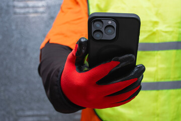 Construction worker wearing protective gloves and reflective safety vest while holding a smartphone. Concept of modern industrial communication, safety at work, and digital technology in construction.