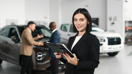 A confident woman in a black suit stands in a car dealership with a tablet in her hands, while a customer and a car dealer negotiate in the background, demonstrating professional experience in car buy