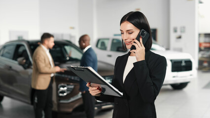 A woman in a business suit talks on the phone while looking through documents, surrounded by cars in a modern car dealership, illustrating the car buying process.