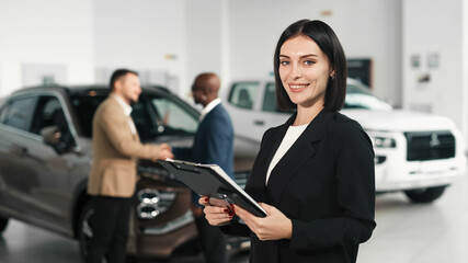 Confident woman in a black suit holding a clipboard stands in a car dealership, with customers...