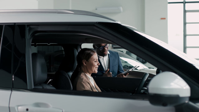 Woman sitting in a car at a dealership, smiling while discussing options with a salesman, showcasing the car buying experience in a modern showroom environment