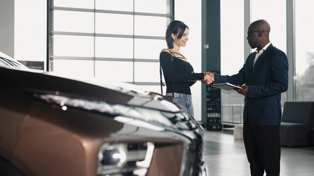 African American man in a suit shakes hands with a Caucasian woman in a car dealership, finalizing the purchase of a new vehicle, showcasing a successful transaction
