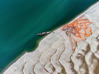 Aerial view of a fishing boat anchored by a sandy shore with nets drying under the sun, creating a striking contrast of teal water and beige sand, Rajshahi, Rajshahi Division, Bangladesh.