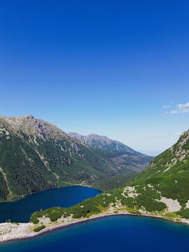 Aerial view of the deep blue lake nestled among the towering mountains, framed by lush green forests under a vast, clear sky, Morskie Oko, Lesser Poland Voivodeship, Poland.