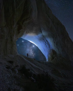 View of a lone figure silhouetted against the ethereal glow of the Milky Way, framed by the rugged arch of a cave at night, Say-Utes, Mangystau Region, Kazakhstan.