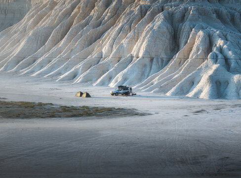 View of an isolated car with a tent on top parked in a vast, desolate landscape of weathered rock formations under a soft, diffused light, Say-Utes, Mangystau Region, Kazakhstan.