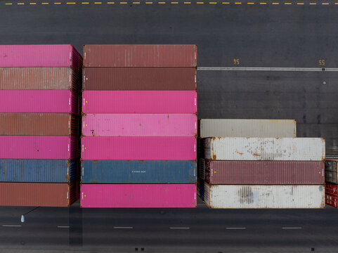 Aerial view of stacked shipping containers painted in vibrant pinks, blues, and browns contrasting against the stark grey asphalt, Berlin, Germany.