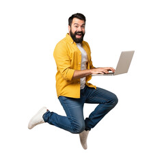 Energetic bearded man in yellow shirt and jeans jumping with laptop isolated on transparent background