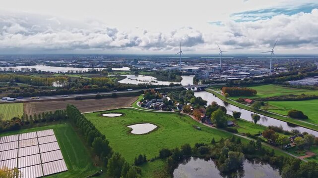 Lush green fields stretch across the landscape interspersed with winding rivers and reflective water bodies Wind turbines stand tall against a backdrop of rolling clouds and distant cityscape