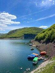 Naklejka premium Scenic view of Uvac Lake in Serbia with colorful boats docked along the shore, surrounded by lush green hills and clear blue sky.