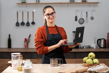 Woman with laptop online in home searching recipe for baking pie with video. Housewife reading recipe in kitchen for cooking