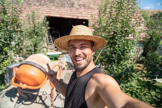 Cheerful construction man in straw hat taking smiling selfie next to cement mixer in rural setting