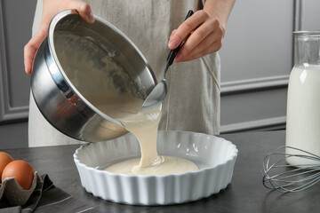 Woman pouring liquid dough from bowl into baking dish at grey table in kitchen, closeup