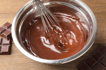 Melted chocolate with whisk in bowl and pieces on wooden table, closeup