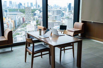 高層ビルを背景に、テーブルの上にノートパソコンとコーヒーカップが置かれたオフィス・カフェ風の空間