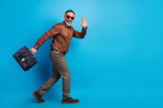 Energetic mature businessman in stylish brown shirt khaki pants and red sunglasses waves hello while carrying a briefcase against a bright blue studio backdrop
