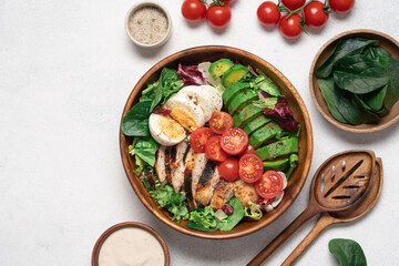Healthy Cobb salad with grilled chicken breast, avocado, cherry tomatoes, lettuce and boiled egg in wooden bowl, top view on light background