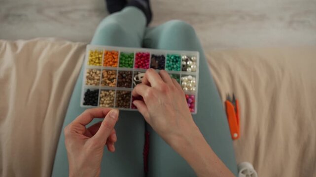 First-person view of a woman's hands skillfully threading colorful beads onto a string, creating a handmade bracelet with a diverse assortment of jewelry making supplies from a craft box