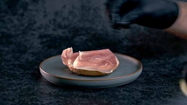 Male hand in black glove assembling delicious bagel sandwich on teal plate. Layers of ham, avocado, pickles, and egg topped with sesame and spices. Dark background,  food preparation.