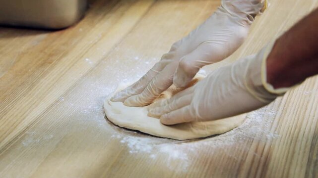 Close-up of gloved hands pressing and shaping dough on a floured wooden surface. Ideal for baking, pizza making, or food preparation themes.