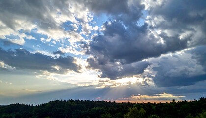 Dramatic skyscape captures sunlight piercing through dense clouds above a dark, tree-covered landscape. Sunbeams radiate brightly